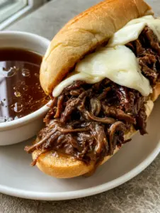 Close-up of a handheld French Dip sandwich showing glistening shredded beef and bubbly melted provolone. (Slow Cooker French Dip Sandwiches)